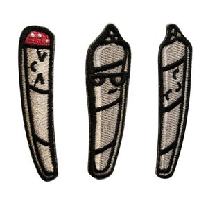 May include: Three embroidered patches in the shape of a marijuana joint. The patches are gray with black outlines. One has a red tip and the letters "VCA". Another has sunglasses and the third has a smiling face.