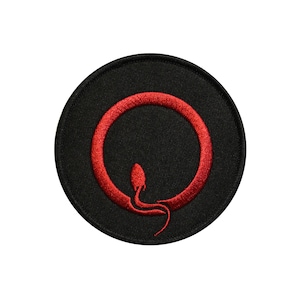 May include: A circular embroidered patch featuring a black background with a vibrant red stitched design. The design consists of a solid red circle with a small, stylized red creature resembling a mouse or rat integrated into the bottom curve.