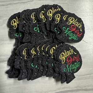May include: A pile of black fabric patches with an embroidered silhouette of a Black woman's head and the text "Black Girl Magic" in gold, green, and red lettering.
