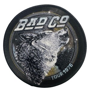May include: Round black patch featuring a howling wolf illustration. The text "BAD CO" is in silver block letters above the wolf. The words "TOUR 1976" are in silver text along the bottom edge of the patch. The wolf is gray and white.