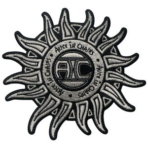 May include: Embroidered patch featuring the Alice in Chains logo within a sunburst design. The patch is primarily black and gray, with the band's name encircling the central logo. The sunburst has pointed rays extending outwards.