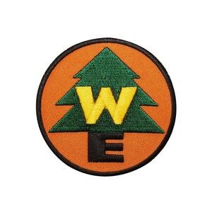 May include: A circular embroidered patch featuring an orange background with a dark green pine tree silhouette in the center. The yellow letter W is stitched over the tree, and the black letter E is positioned below the tree on the orange field.