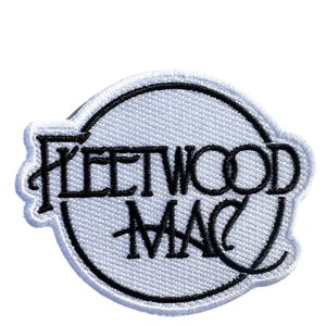 May include: A circular embroidered patch featuring a white background with a black border and the text Fleetwood Mac in black stylized lettering. The design is stitched with a visible thread texture throughout the entire circular shape.