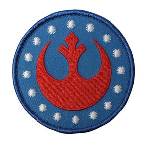 May include: A circular patch with a blue background and a red emblem. The emblem is a stylized crescent shape with a central design. White star-like shapes are evenly spaced around the emblem's perimeter. The patch has a blue border.