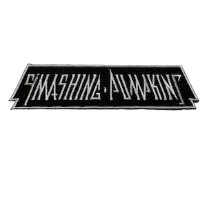 May include: A black and white embroidered patch featuring the text "SMASHING PUMPKINS" in a stylized font. The patch has a rectangular shape with angled edges and a white border.