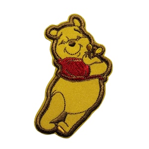 May include: Embroidered patch featuring a smiling yellow bear with a red shirt and brown outline. The bear is holding a small object, possibly honey. The patch is designed for application on clothing or accessories.