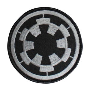 May include: Round black patch with a white and gray Imperial symbol. The symbol is a stylized cogwheel design with eight spokes. The patch has a black border and is likely an iron-on or sew-on patch.