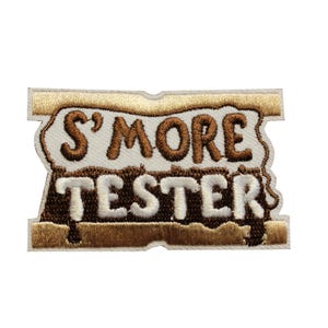 May include: Embroidered patch with the words "S'MORE TESTER" in brown and white. The patch has a light brown border and a design that resembles a s'more. Perfect for jackets, bags, or any fabric item.