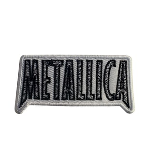 May include: A rectangular patch with the word "METALLICA" embroidered in black on a gray background. The patch has a white border and a slightly curved shape, with the text in a bold, stylized font.