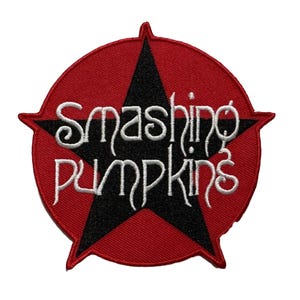 May include: A red and black embroidered patch featuring the text "Smashing Pumpkins" in white script. The text is centered over a black starburst design, set against a red circular background. The patch has a textured appearance.