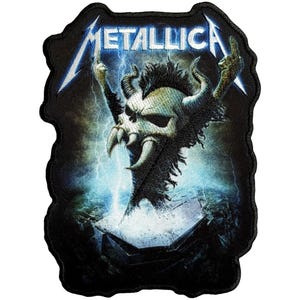 May include: Embroidered patch featuring the band name "METALLICA" in white and blue, above a skull-like creature with horns and raised hands. The design includes lightning bolts and a stormy background, all on a black patch.