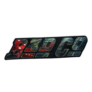 May include: A rectangular patch with the text "BAD CO" in a stylized font. The letters are filled with a colorful pattern of red, blue, and yellow. The patch has a black border.