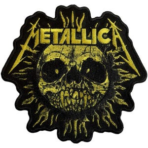 May include: Black and yellow embroidered patch featuring the word "METALLICA" above a skull design. The skull is surrounded by a sunburst pattern. The patch has a jagged edge.