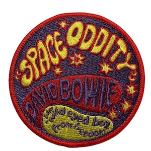 May include: A circular embroidered patch with a red border and a purple background. The patch features the words "SPACE ODDITY" in yellow, arched across the top, and "DAVID BOWIE" in red. Below, it reads "Star eyed boy from freedom" in yellow.