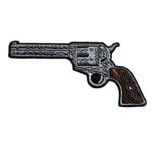 May include: Embroidered patch of a revolver with intricate detailing. The gun features a silver barrel and body with a brown, textured handle. The patch is designed with a black border and is likely for application on clothing or accessories.