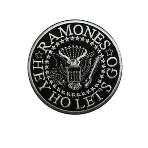May include: A round, black and white embroidered patch featuring the words "THE RAMONES" and "HEY HO LET'S GO" around a central emblem. The emblem includes an eagle, stars, and a banner.