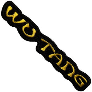 May include: Black embroidered patch with the word "WAKE" in yellow, stylized lettering. The patch has a unique, elongated shape. The yellow thread provides a striking contrast against the black background.