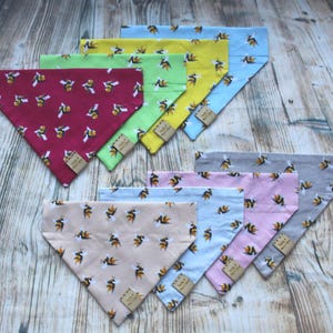 May include: Assortment of ten colorful dog bandanas with a bee pattern. The bandanas are made of cotton fabric and feature a variety of colors, including red, pink, blue, green, yellow, and gray. Each bandana has a small brown tag with the text "Made with Love" printed on it.
