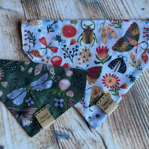 May include: Two triangular pet bandanas featuring insect and floral patterns. One bandana has a white background with colorful beetles, moths, and flowers. The other is dark green with light blue dragonflies and butterflies. Both have tan labels reading Teddy & Max's Spot.