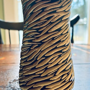 May include: A handcrafted ceramic vase with a textured surface. The vase features a repeating pattern of raised, curved lines in a light brown color against a dark brown background. The vase is cylindrical with a slightly flared top.