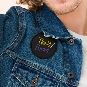 May include: A person wearing a blue denim jacket with a black button with the text "They/Them" in yellow and purple lettering.