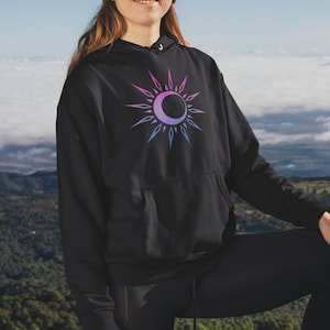 May include: A person wearing a black hoodie with a pink and blue sun and moon graphic, a black beanie, and sunglasses, standing on a rocky mountaintop with a golden retriever dog.
