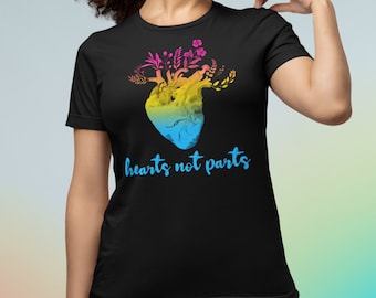 May include: Black t-shirt featuring a colorful heart design with floral accents and the text "hearts not parts" in blue. The heart design transitions from pink to yellow to blue. The shirt is worn with blue jeans.
