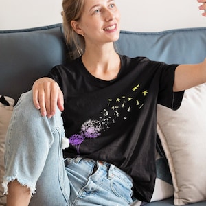 May include: A woman wearing a black t-shirt with a dandelion graphic and yellow birds printed on it. She is sitting on a blue couch and taking a selfie with a white smartphone.