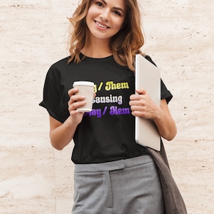May include: A black t-shirt with the text "They/Them causing May/Them" in yellow and purple gradient lettering.