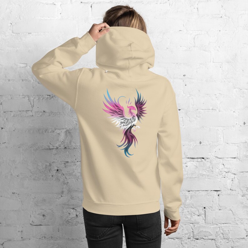 May include: Beige hoodie with a colorful phoenix design on the back. The phoenix is depicted with shades of pink, purple, blue, and white. The hoodie has a hood and long sleeves.