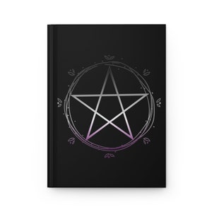Puede incluir: Una libreta negra con un pent&aacute;culo plateado rodeado de un c&iacute;rculo de hojas. El pent&aacute;culo es una estrella de cinco puntas con un degradado de morado y blanco.