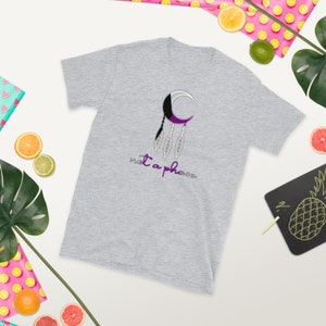 以下が含まれることがあります： グレーのTシャツに、紫と黒の三日月型のドリームキャッチャーのデザインと、紫色の「not a phase」という文字が書かれています。
