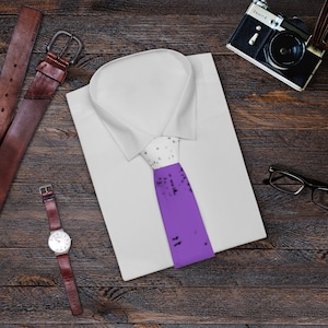 May include: A white dress shirt with a purple tie with black speckles. The tie is on a wooden surface with a brown leather belt, a watch, and a camera.