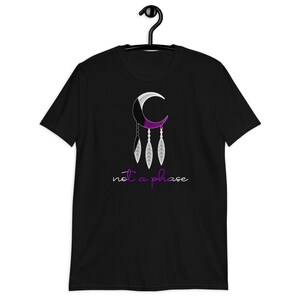 以下が含まれることがあります： 三日月と羽根が描かれた黒いTシャツ。三日月は紫色と白で、羽根は三日月からぶら下がっている。三日月の下に「not a phase」という文字が書かれている。