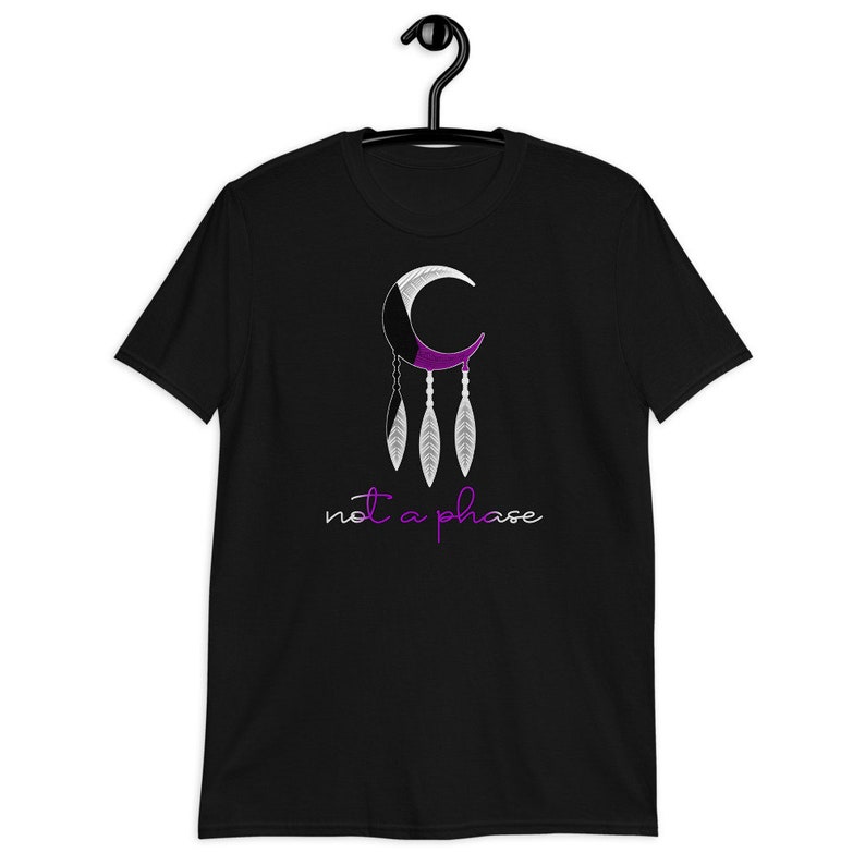 以下が含まれることがあります： 三日月と羽根が描かれた黒いTシャツ。三日月は紫色と白で、羽根は三日月からぶら下がっている。三日月の下に「not a phase」という文字が書かれている。