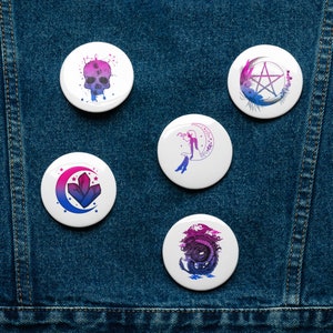 May include: Five round, white button pins with different designs. Each pin features a watercolor-style design in shades of pink, blue, and purple. The designs include a skull, a crescent moon with a fairy, a pentagram, a crystal, and a dragon.