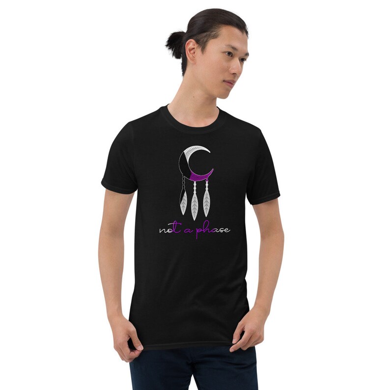 以下が含まれることがあります： 三日月と羽根が描かれた黒いTシャツ。三日月は紫色と白で、羽根は三日月からぶら下がっている。三日月の下に「not a phase」という文字が書かれている。