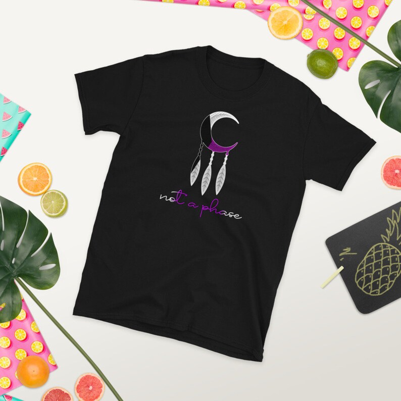以下が含まれることがあります： 紫と白の三日月と羽根が描かれた黒いTシャツで、紫色の「not a phase」という文字が書かれています。