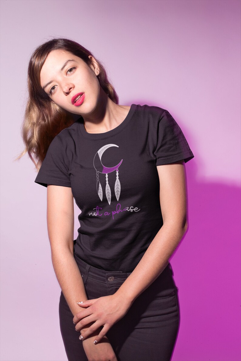 以下が含まれることがあります： 紫と白の三日月と羽根のグラフィックが描かれた黒いTシャツ。グラフィックの下に「not a phase」という文字が紫で印刷されています。