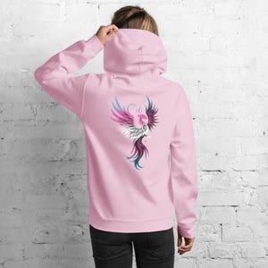 May include: A light pink hoodie with a colorful phoenix design on the back. The phoenix is depicted with shades of pink, blue, white, and black. The hoodie has a hood and long sleeves.