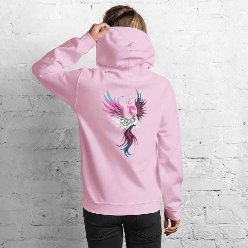 May include: A light pink hoodie with a colorful phoenix design on the back. The phoenix is depicted with shades of pink, blue, white, and black. The hoodie has a hood and long sleeves.