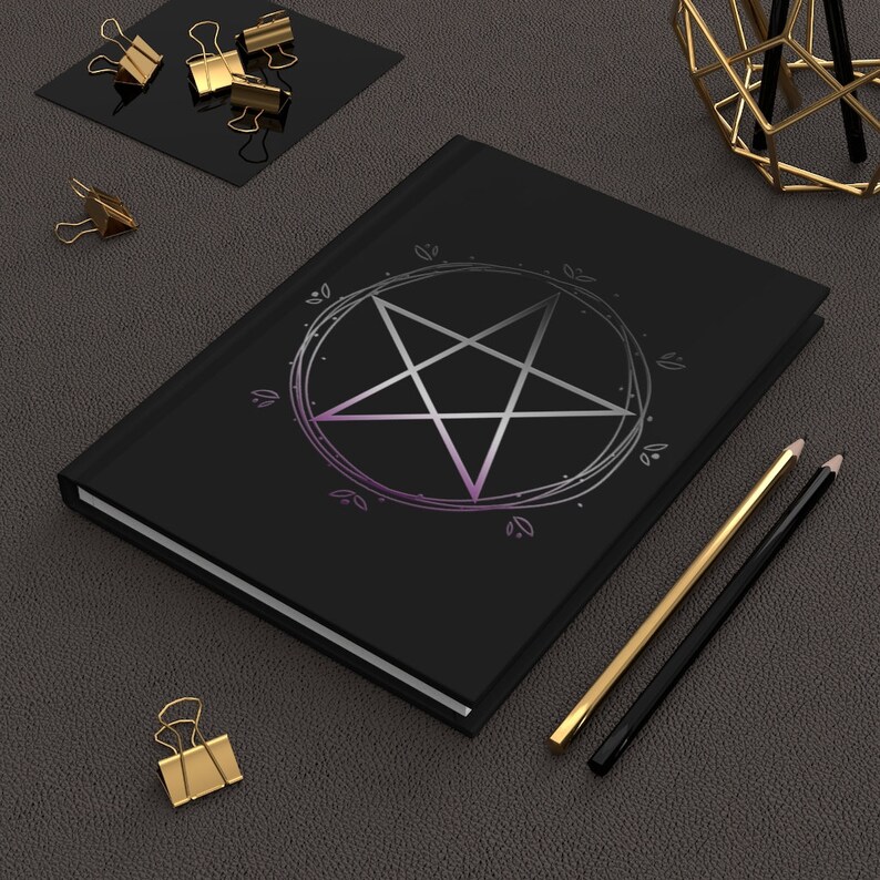 Puede incluir: Un cuaderno negro con un pentagrama plateado dentro de un c&iacute;rculo con un degradado morado. El cuaderno est&aacute; sobre una superficie gris texturizada con dos l&aacute;pices y clips de papel dorados.