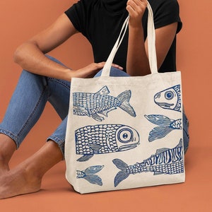 May include: A white canvas tote bag with a blue fish design. The bag features four fish, each with a unique pattern and design. The fish are all facing different directions. The bag is being held by a person wearing a black shirt and blue jeans.