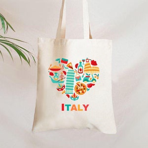 Op de afbeelding: Cr&egrave;mekleurige draagtas met een hartvormig ontwerp gevuld met Italiaanse iconen zoals de scheve toren van Pisa, pizza en het Colosseum. Het woord "ITALY" is in het rood gedrukt. De tas wordt door een hand vastgehouden.