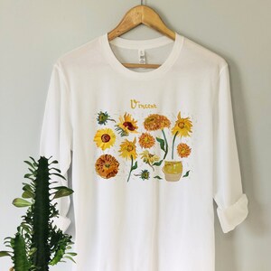 May include: White long-sleeve t-shirt with a graphic print of sunflowers in a vase. The text "Vincent" is printed on the shirt.