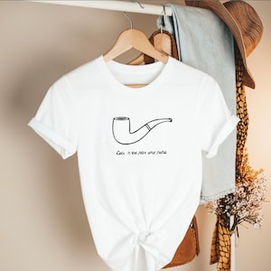 May include: White t-shirt with a black outline drawing of a pipe. The text "Ceci n'est pas une pipe" is below the pipe. The shirt is hanging on a wooden hanger with other clothing and a hat in the background.