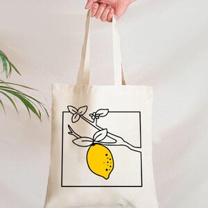 May include: A cream-colored tote bag with a black and yellow lemon illustration. The lemon is on a branch with leaves, enclosed in a black square. The bag is held by a hand.