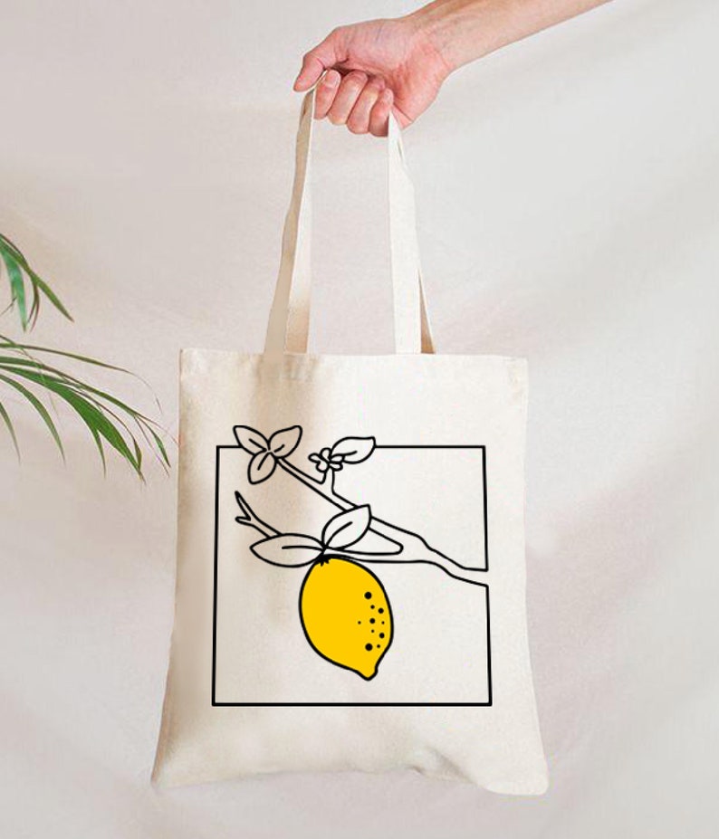 May include: A cream-colored tote bag with a black and yellow lemon illustration. The lemon is on a branch with leaves, enclosed in a black square. The bag is held by a hand.