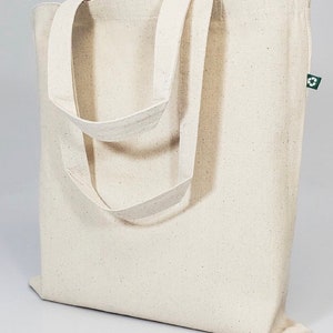 Op de afbeelding: Een lichtbeige canvas draagtas met twee lange handvatten. De tas heeft een eenvoudig ontwerp en is gemaakt van natuurlijk materiaal. Een klein groen label met een recyclingsymbool is aan de zijkant bevestigd.