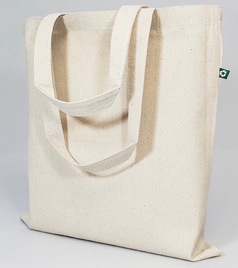 Op de afbeelding: Een lichtbeige canvas draagtas met twee lange handvatten. De tas heeft een eenvoudig ontwerp en is gemaakt van natuurlijk materiaal. Een klein groen label met een recyclingsymbool is aan de zijkant bevestigd.